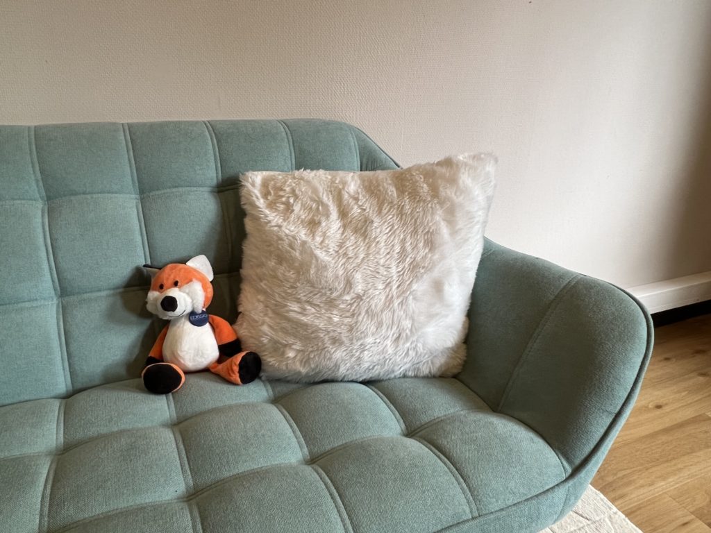 La peluche renard Edifox, confortablement assise sur un canapé contre un coussin.